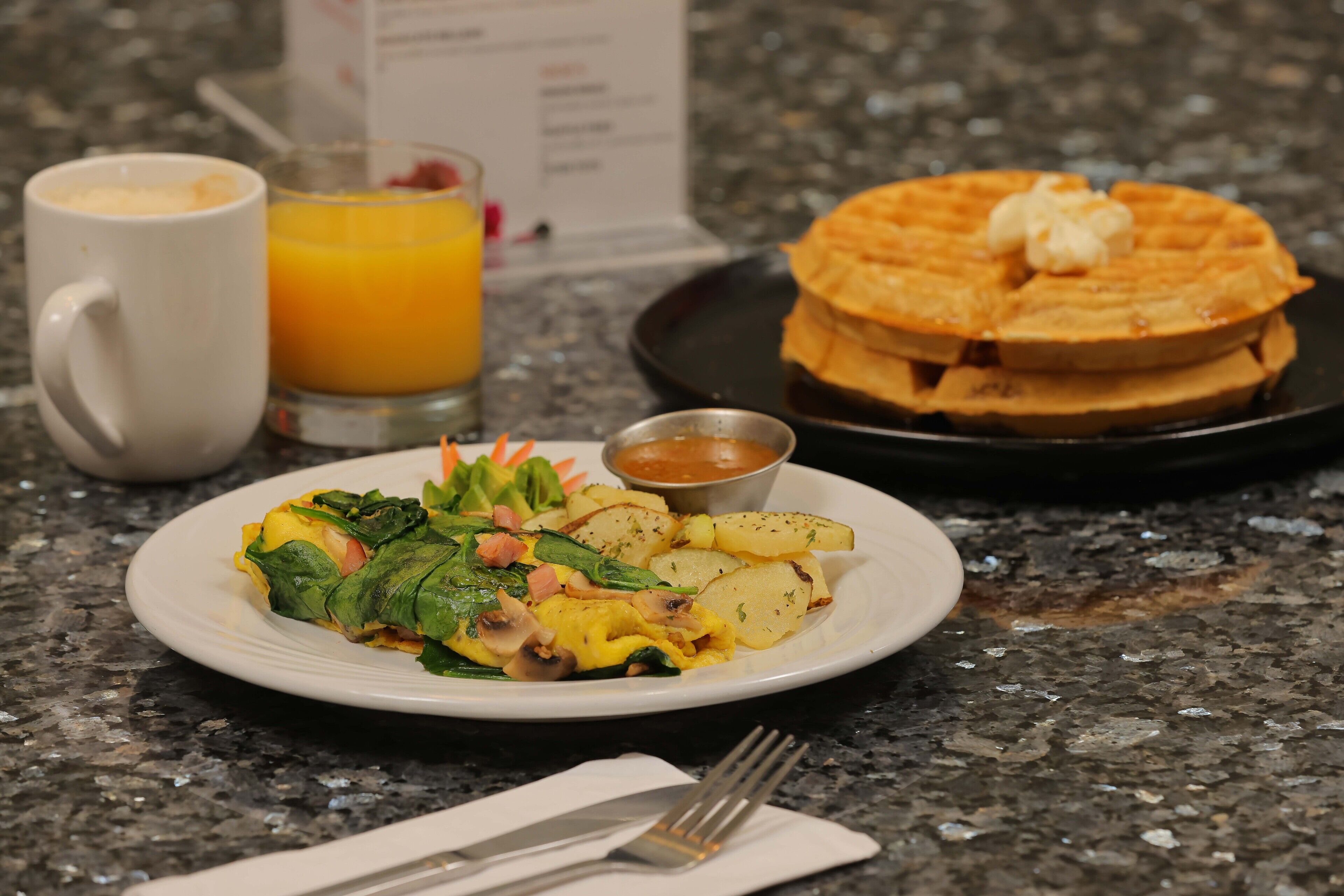 Une assiette de petit-déjeuner avec des œufs, des pommes de terre et des légumes verts, une pile de gaufres, un verre de jus d’orange et une tasse de café.