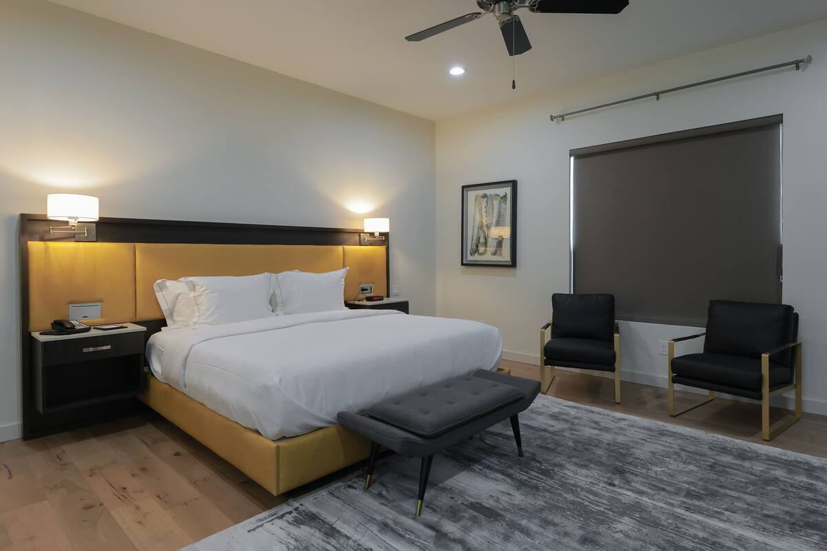 A modern hotel room with a large bed, bedside lamps, a ceiling fan, a desk with a phone, two chairs, and a large window.