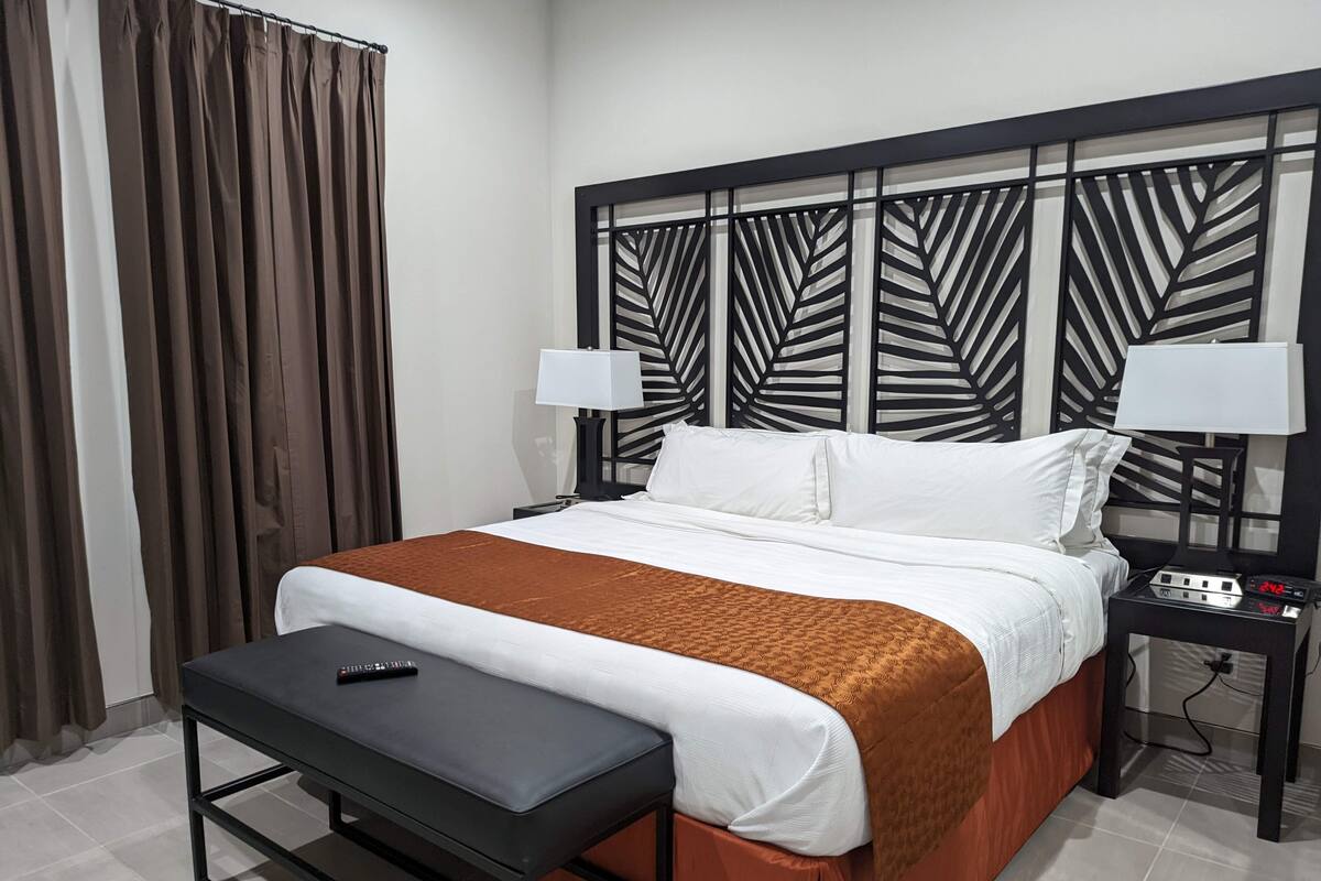 A neatly made bed with a brown blanket, a black bedside table with a remote control, and a nightstand with a lamp and a digital clock.