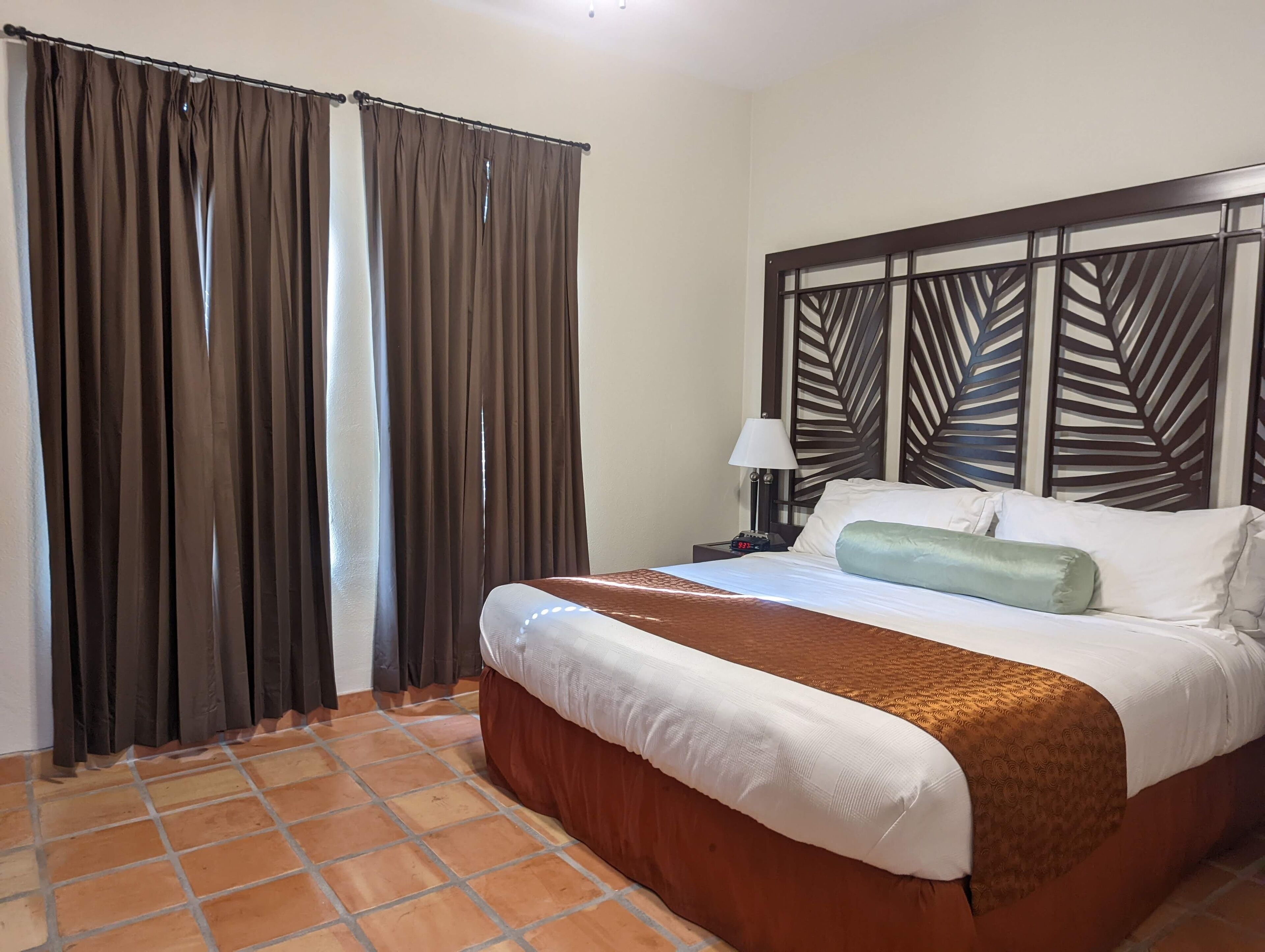A hotel room with a bed, curtains, a nightstand with a lamp, and a decorative headboard.