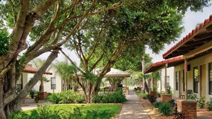 A pathway flanked by lush greenery and trees, leading to a building with a red-tiled roof and a covered patio area. - The Bungalows on Shary, Mission-McAllen (Mission)