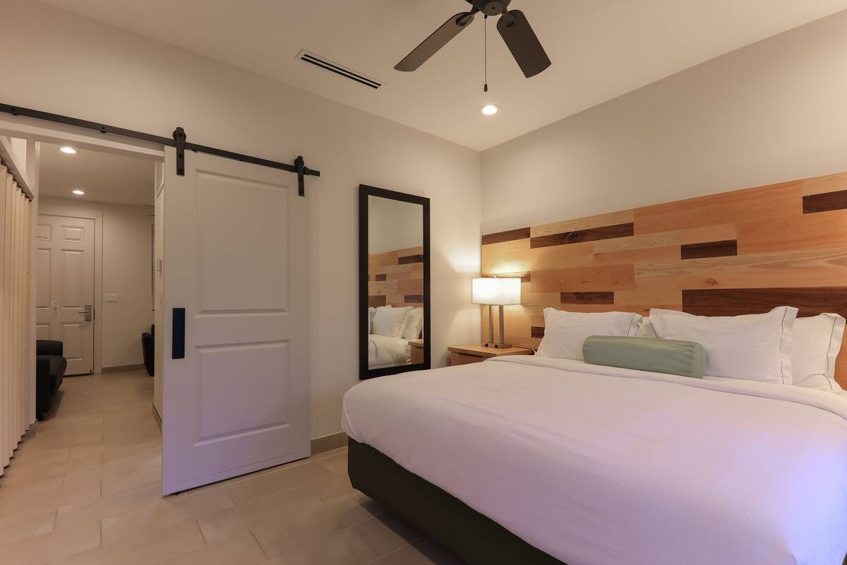 A hotel room with a bed, a nightstand, a mirror, a ceiling fan, and a sliding door leading to another room.