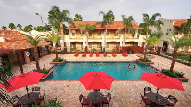 A pool area with red umbrellas, lounge chairs, and a building with a tiled roof. - The Bungalows on Shary, Mission-McAllen (Mission)