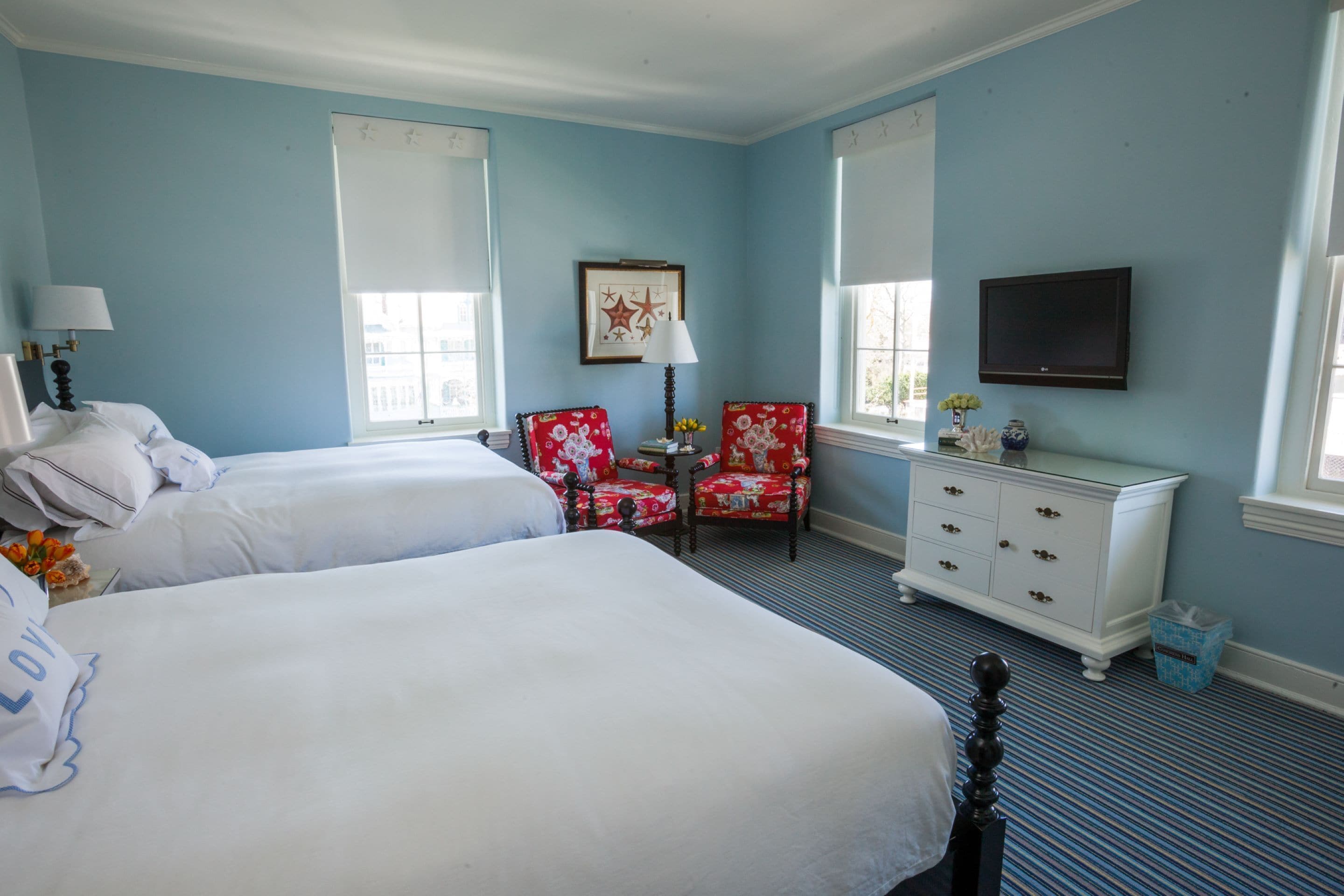 A hotel room with two beds, a red floral armchair, a white dresser, and a television mounted on the wall.