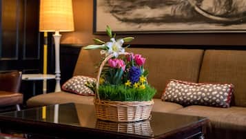 A living room with a wicker basket of flowers on a glass coffee table, a sofa with patterned cushions, a lamp, and a framed painting on the wall.