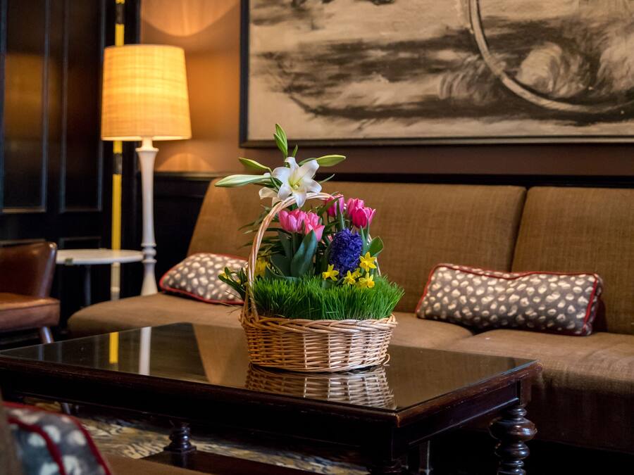 A living room with a wicker basket of flowers on a glass coffee table, a sofa with patterned cushions, a lamp, and a framed painting on the wall.