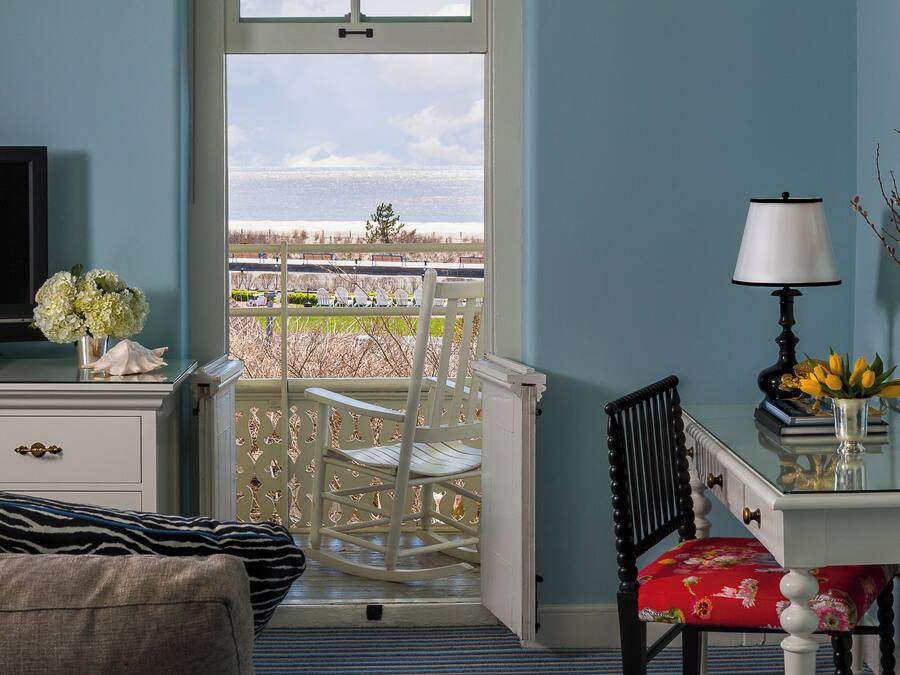 A room with a view of the beach, a rocking chair, a desk with a lamp, and a television.