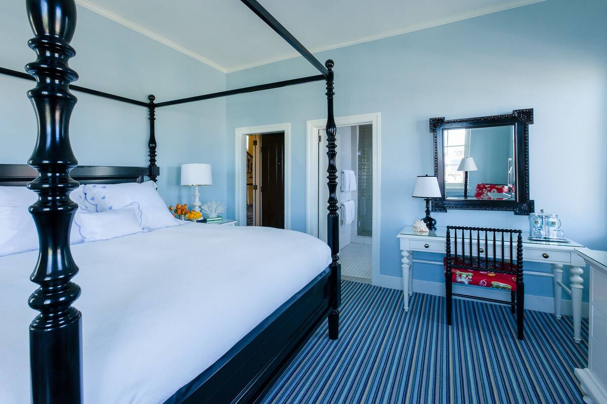 A four-poster bed with white linens, a white dresser with a mirror, and a striped floor.