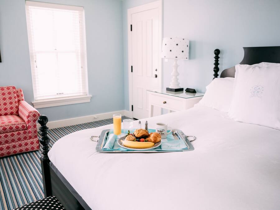 A neatly made bed with a tray of breakfast items, a bedside table with a lamp, and a red patterned chair in a room with light blue walls.