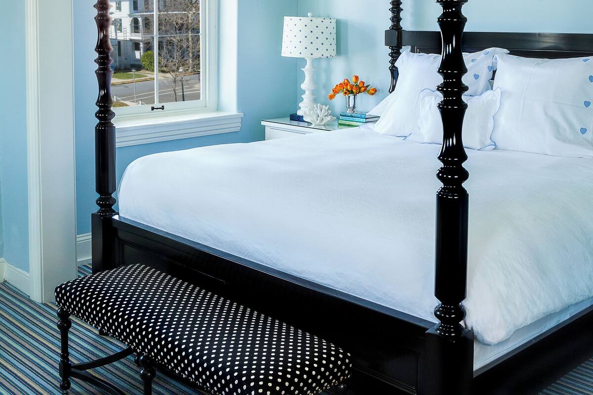 A four-poster bed with a bench, a nightstand with a lamp, and a window with a view of buildings.