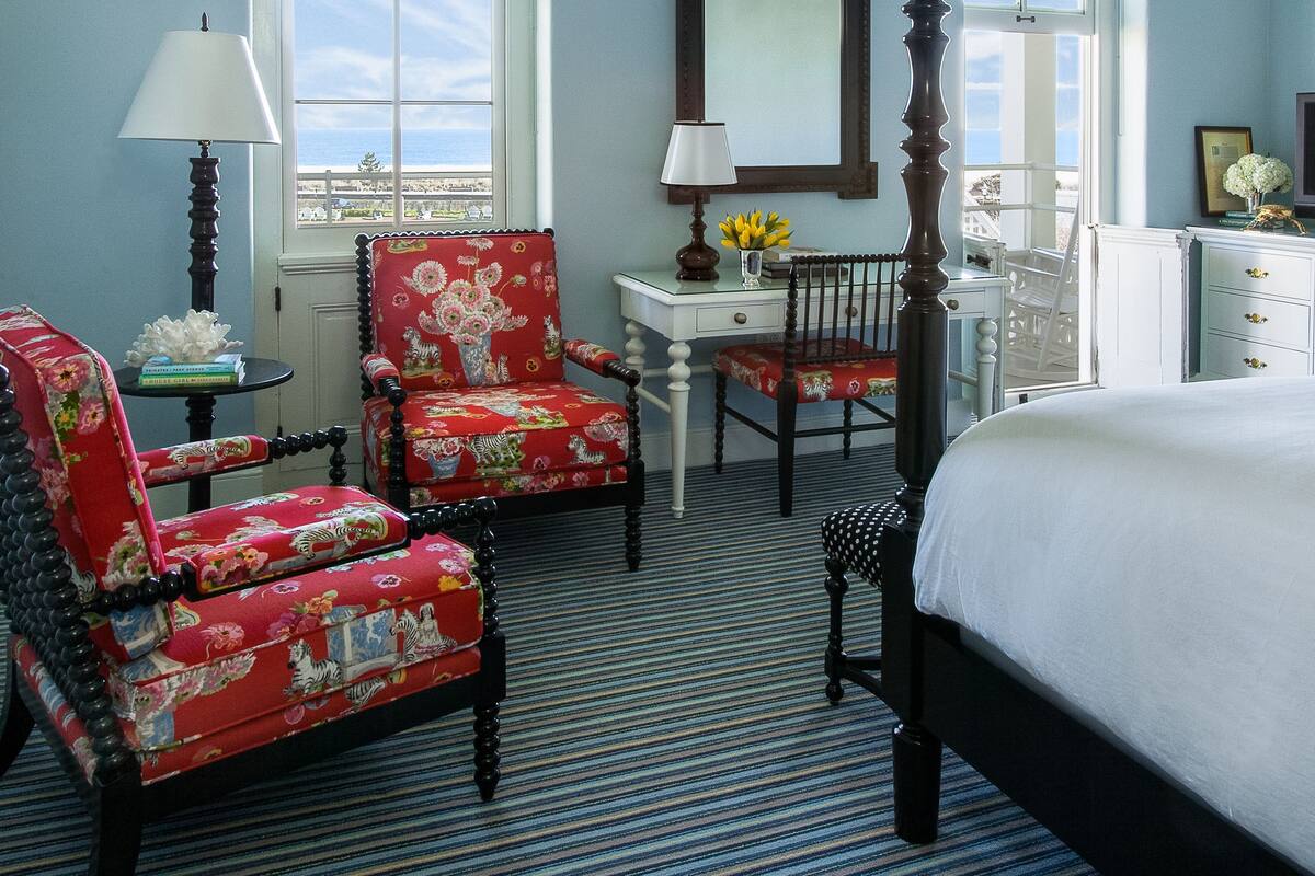 A bedroom with a four-poster bed, two chairs with floral upholstery, a small table with a lamp, and a dresser.
