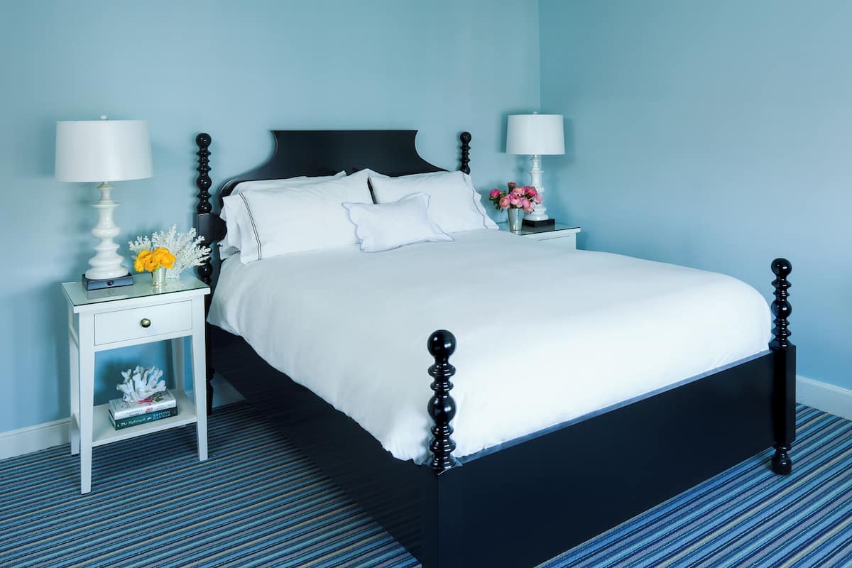 A neatly made bed with white linens, two bedside tables with lamps, a small nightstand with a vase of flowers, and a blue striped carpet.