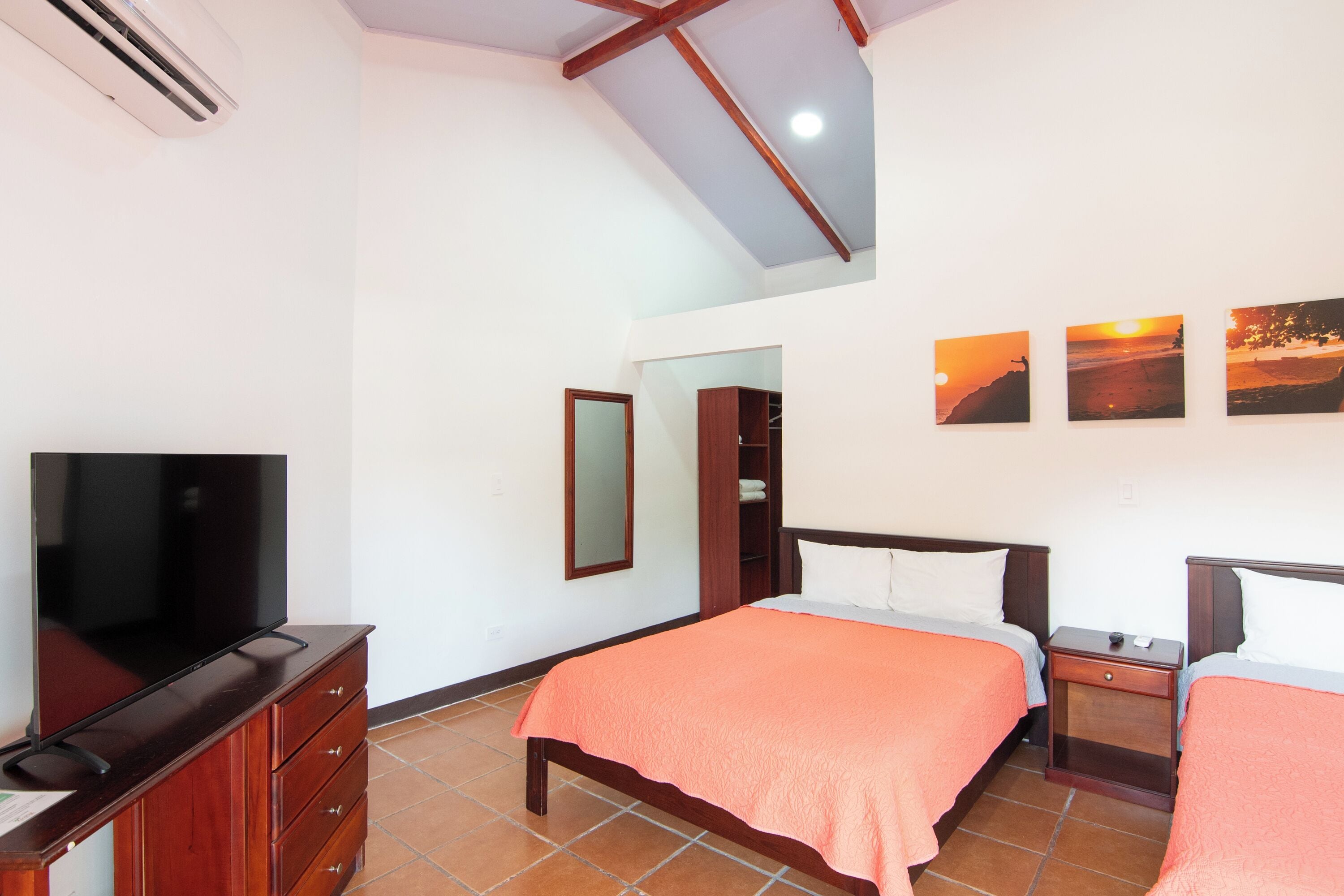 Une chambre avec une télévision, des meubles en bois, un lit recouvert d’une couverture orange et trois tableaux représentant des couchers de soleil accrochés au mur.