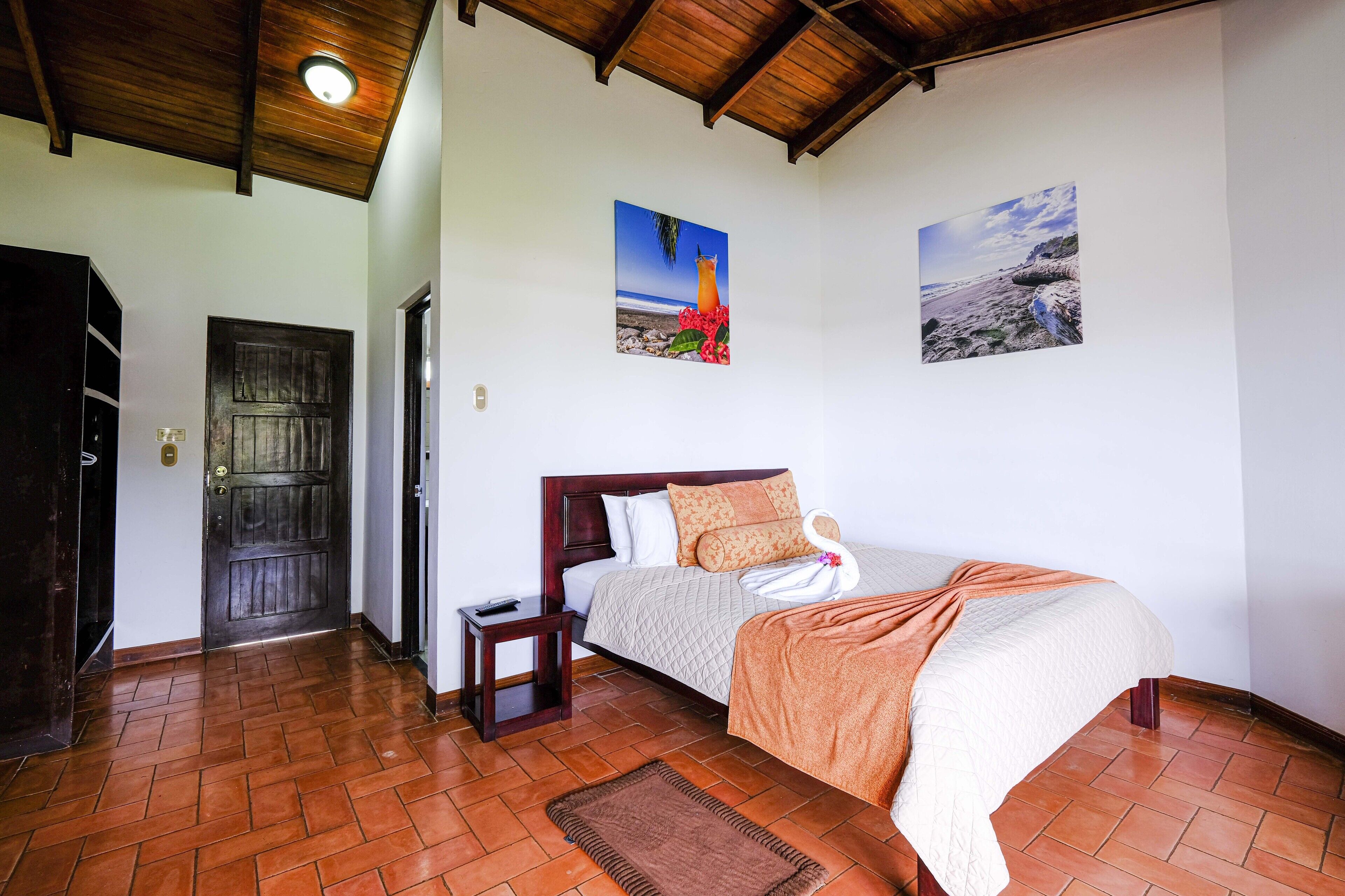 A bedroom with a wooden ceiling, a bed with a white and orange blanket, a small bedside table, and two wall-mounted pictures of landscapes.