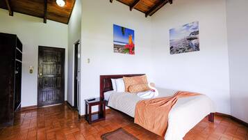 A bedroom with a wooden ceiling, a bed with a white and orange blanket, a small bedside table, and two wall-mounted pictures of landscapes.