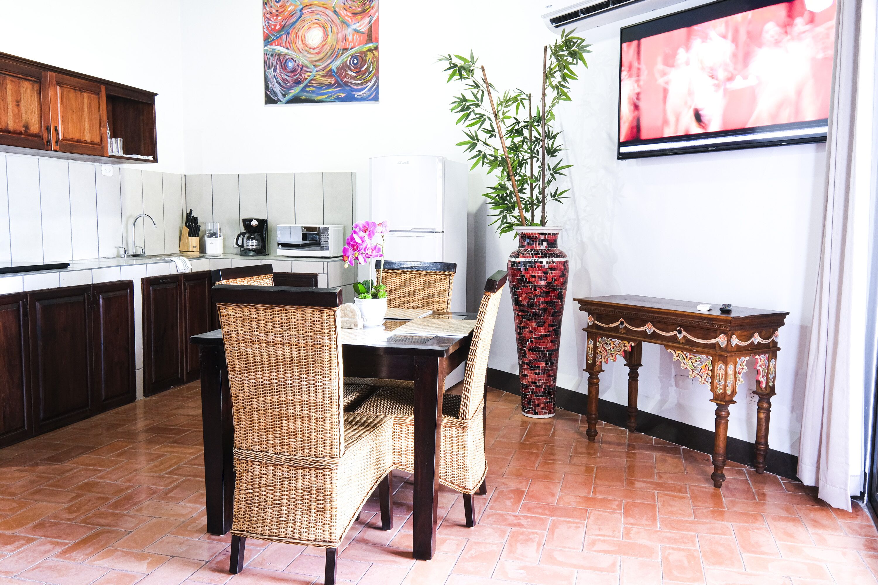 A kitchen with dark wooden cabinets, a white tiled backsplash, a dining table with wicker chairs, a red patterned vase, and a television on the wall.