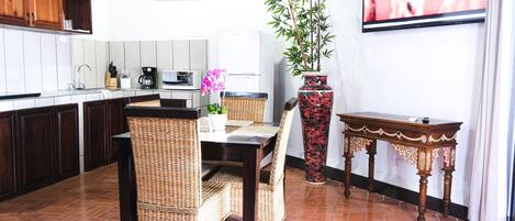 Una cocina con armarios de madera oscura, salpicadero de azulejos blancos, una mesa de comedor con sillas de mimbre, un jarrón rojo estampado y un televisor en la pared.