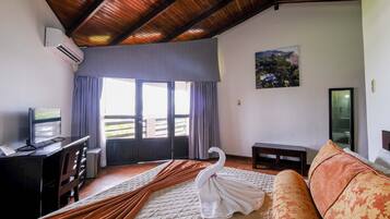 Quarto de hotel com cama, escrivaninha, televisão, janela com cortinas, um quadro na parede e teto de madeira.