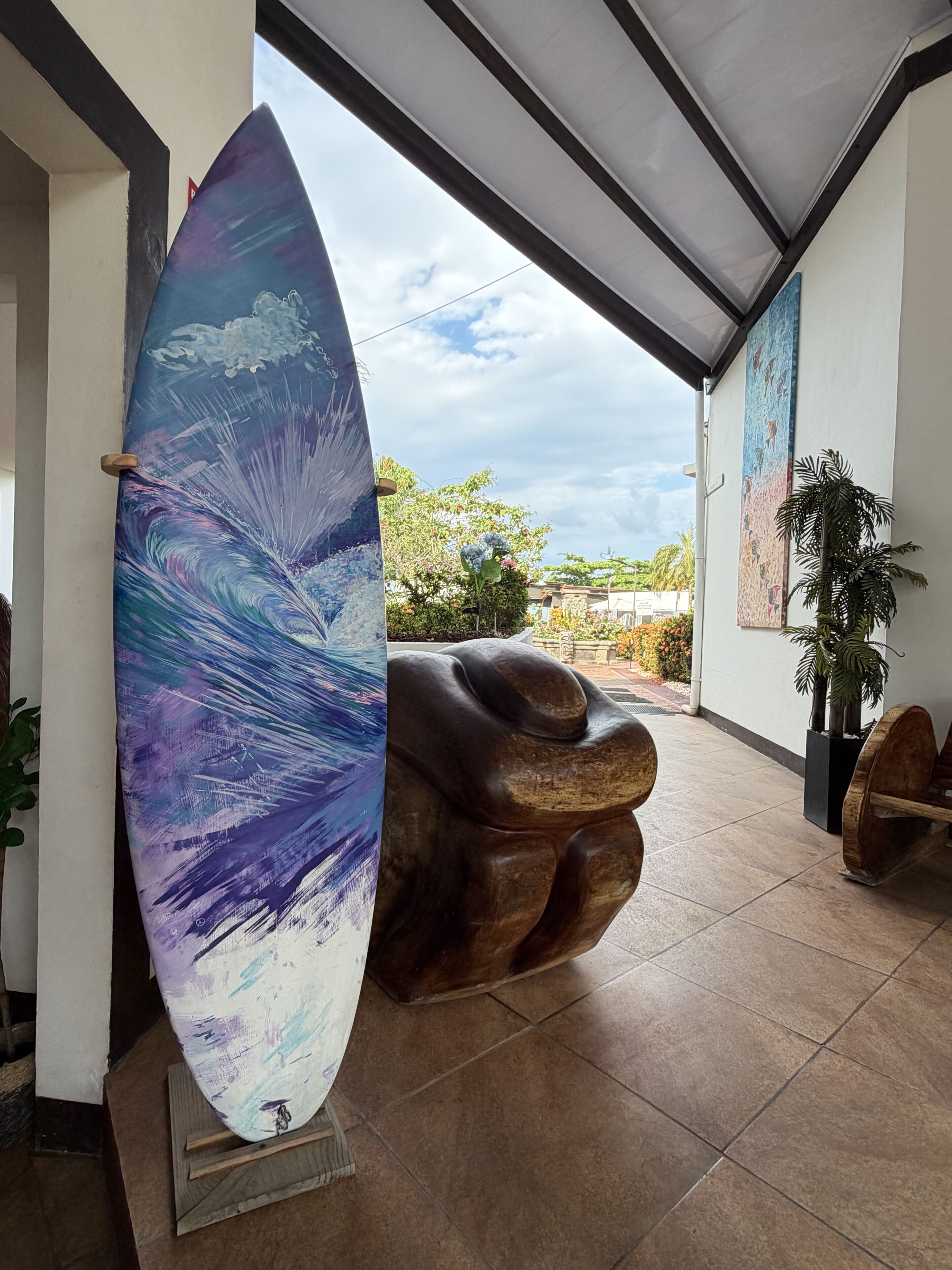 A surfboard leaning against a wall, a wooden sculpture, and a potted plant in a covered area with a view of the outside.