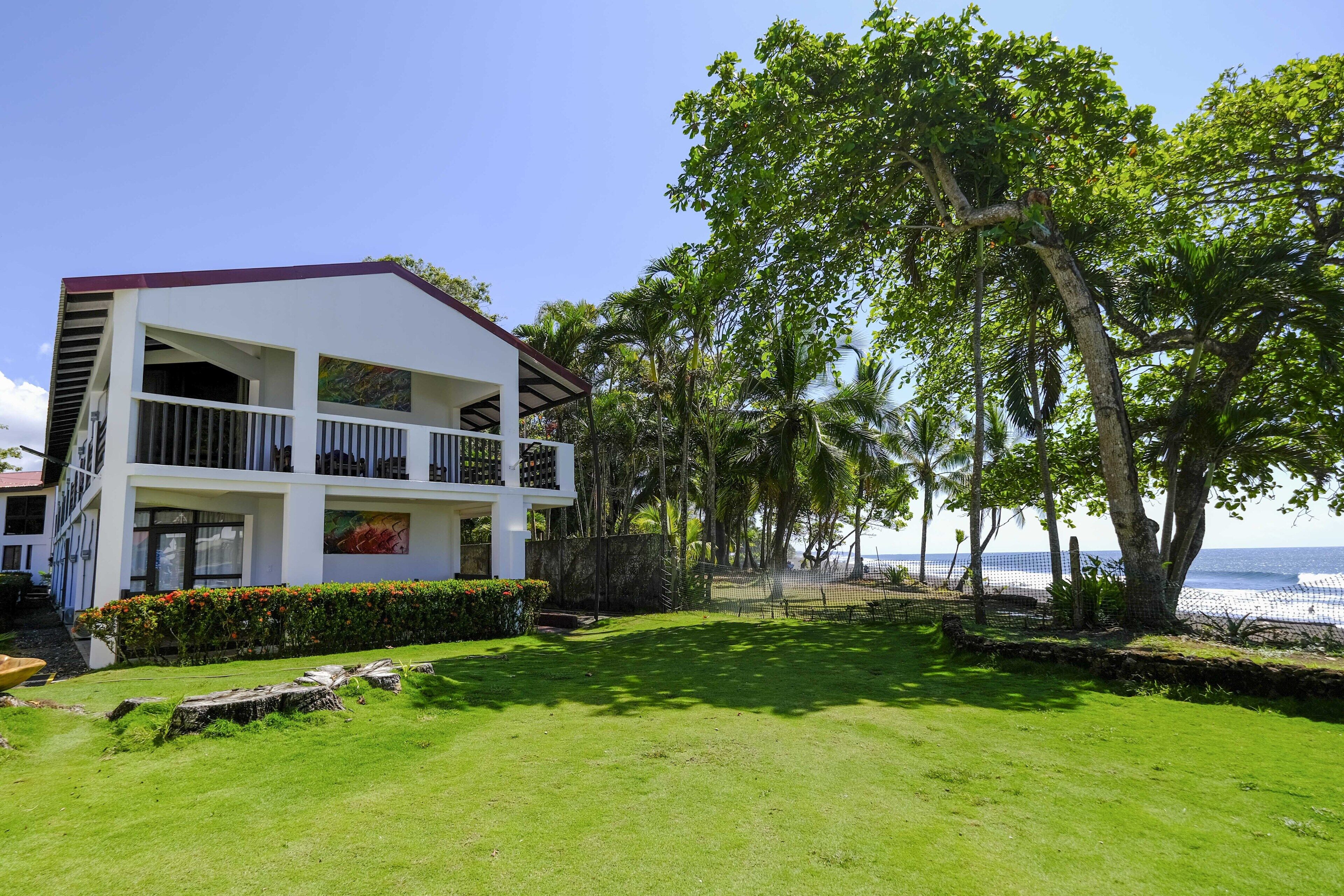 A two-story building with a balcony, surrounded by lush greenery and a view of the beach.
