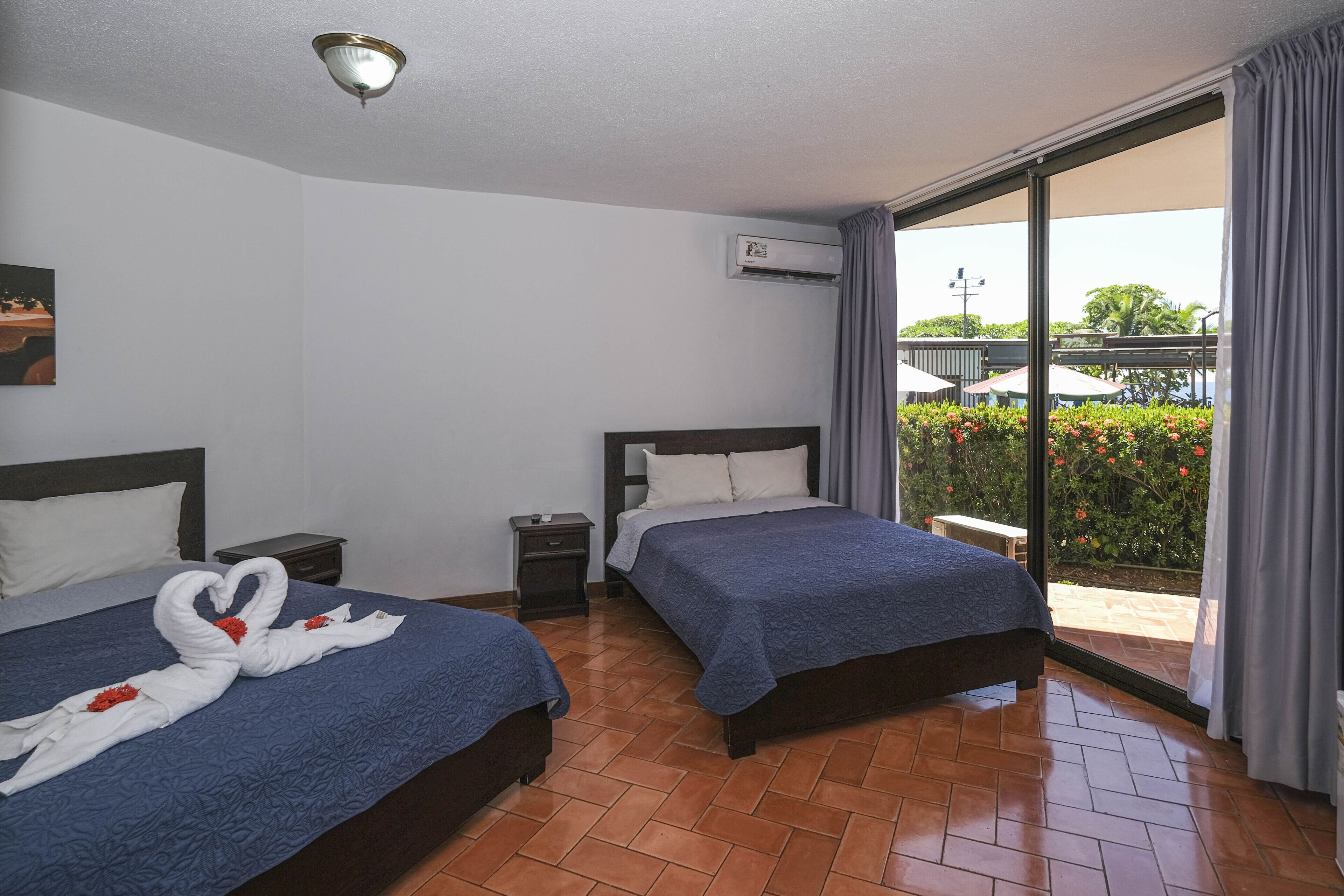 A hotel room with two beds, a nightstand, an air conditioning unit, and a view of the outdoors.