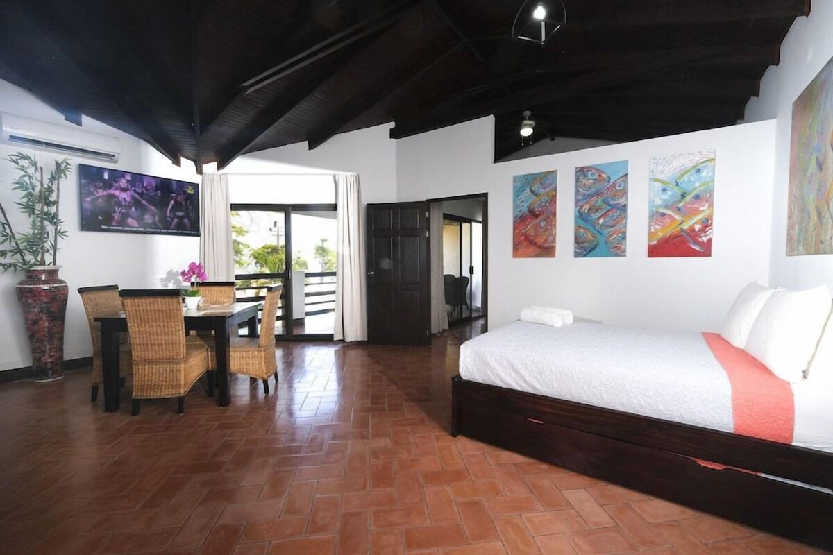 A room with a bed, a dining table with chairs, a TV, and artwork on the walls.