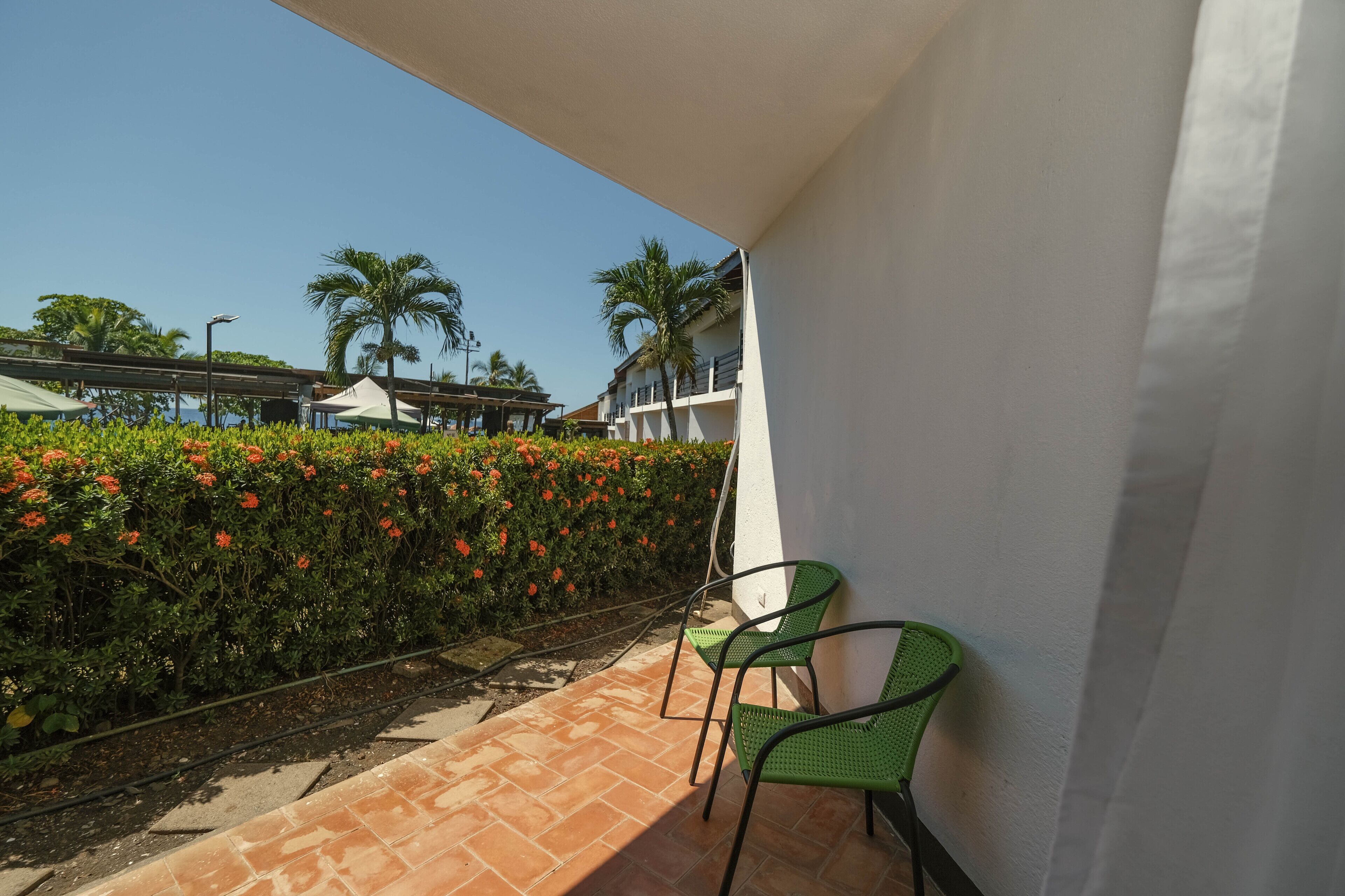 A balcony with two green chairs, a tiled floor, and a hedge with red flowers.
