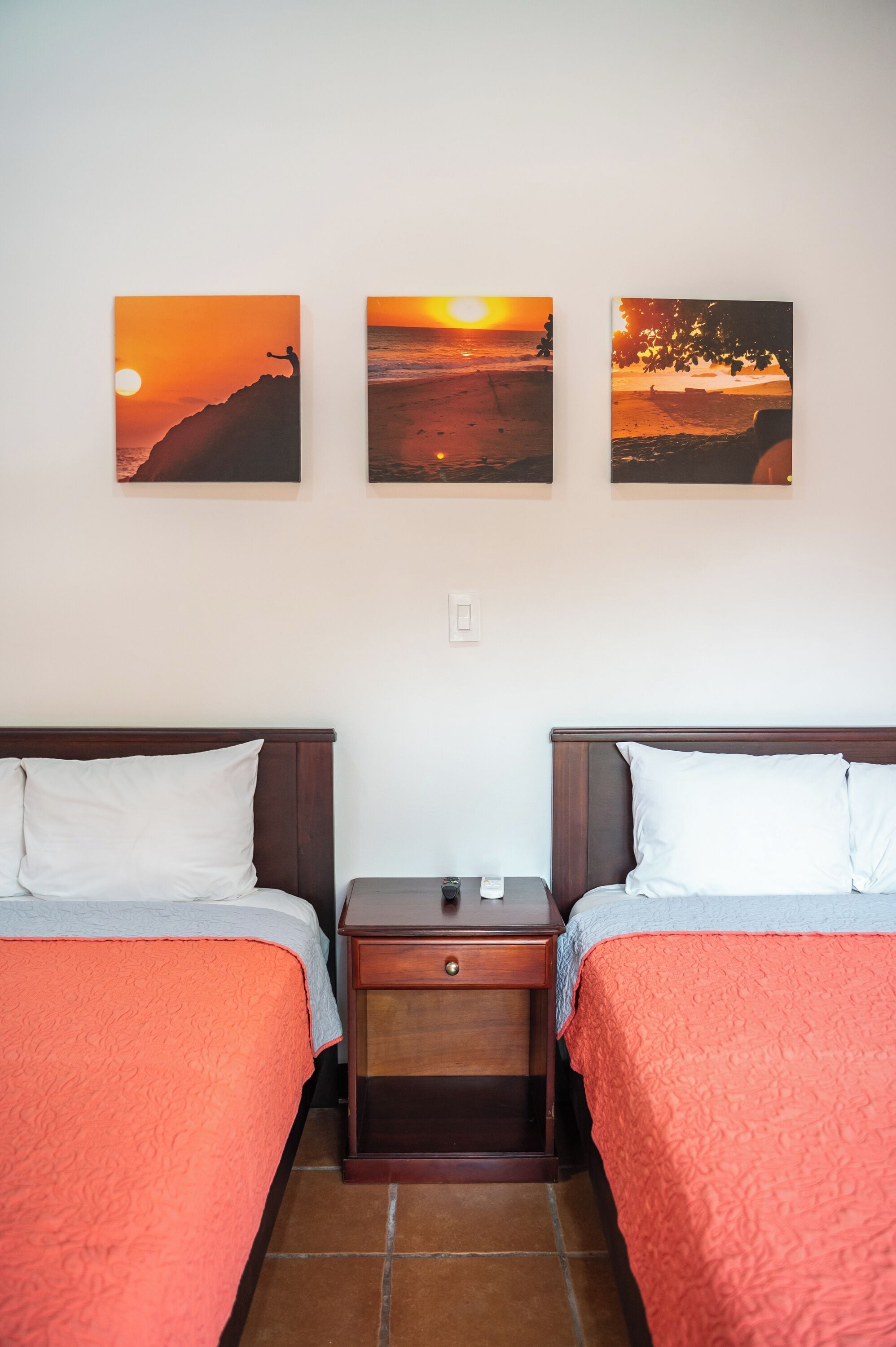 A hotel room with two beds, each with a red bedspread and white pillows. There are three paintings on the wall depicting sunsets.