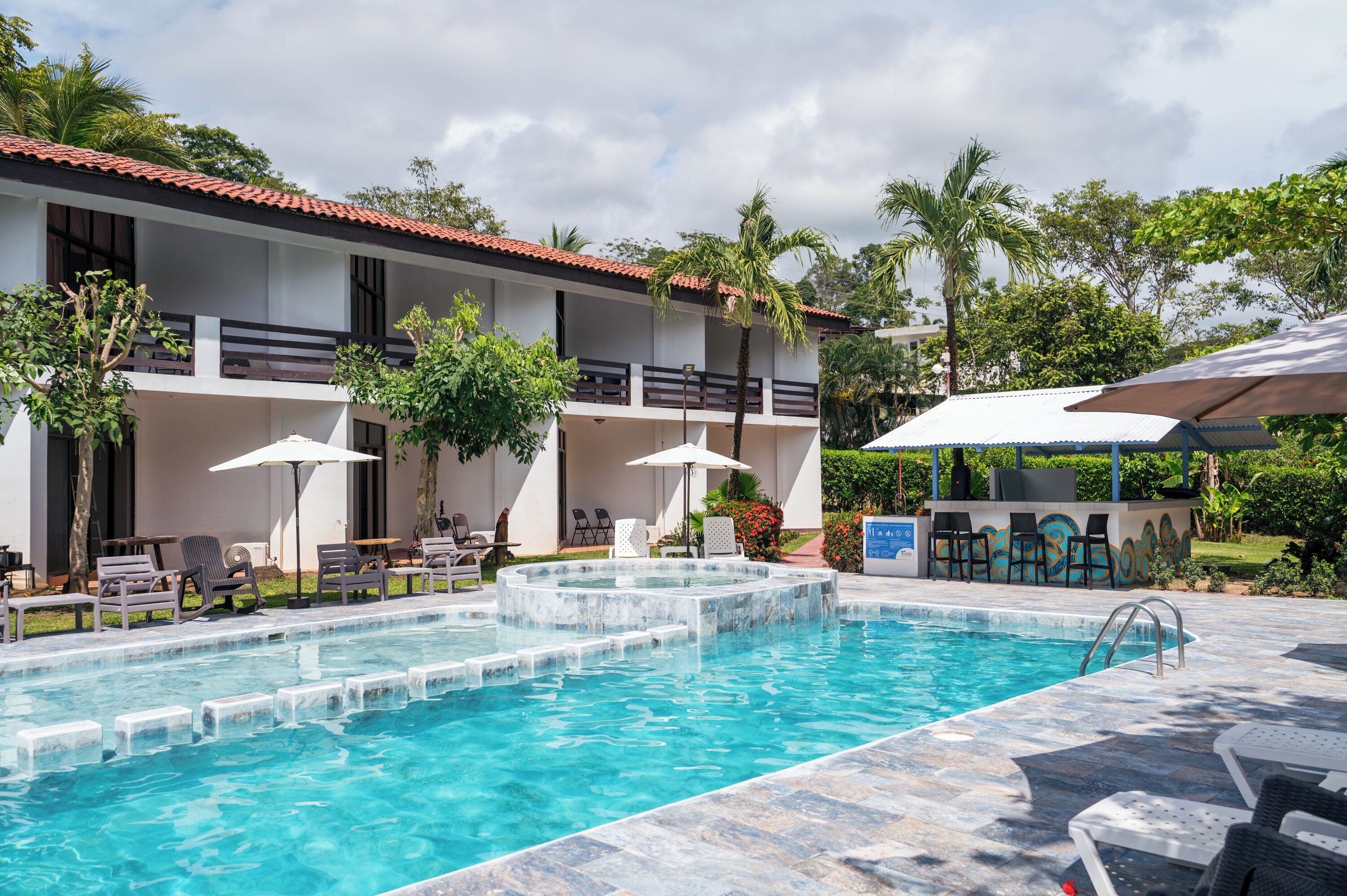 A swimming pool with lounge chairs and umbrellas, surrounded by a building with a tiled roof and balconies.