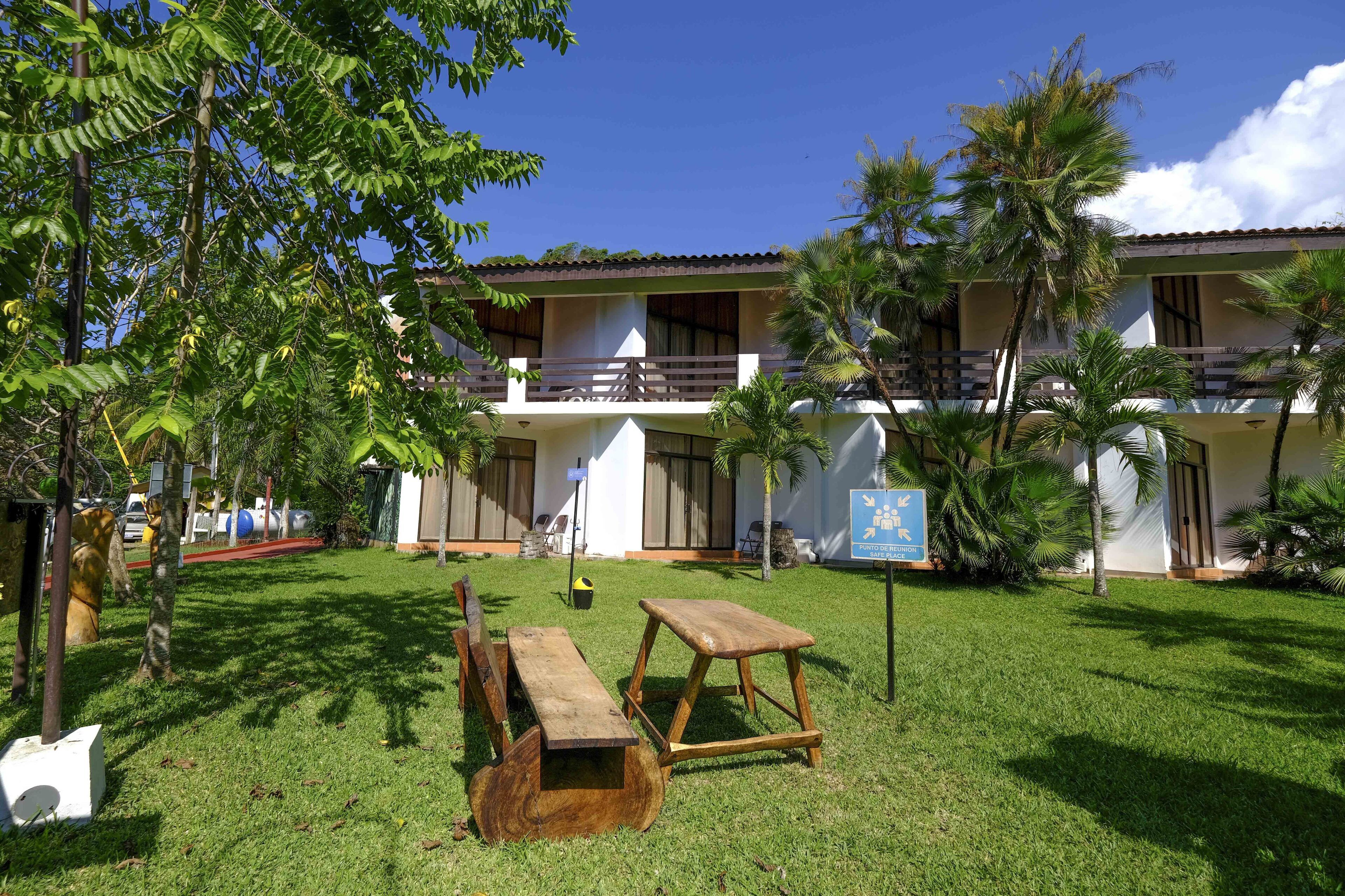 A building with a balcony, surrounded by a grassy area with trees and a wooden bench.