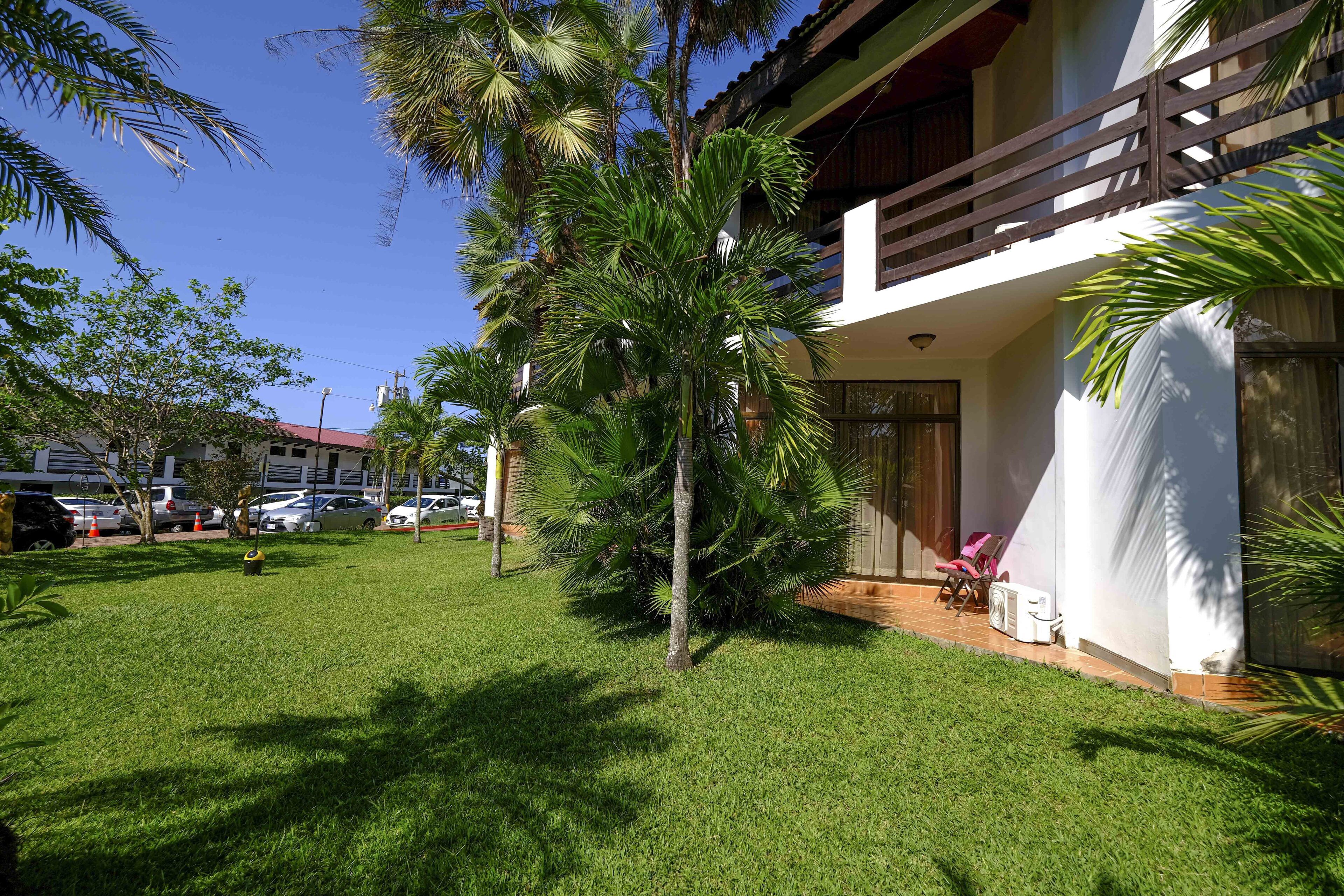 A well-maintained lawn with palm trees, a balcony with a chair, and a building with a red roof in the background.