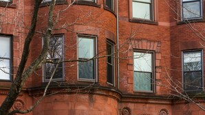 A red brick building with multiple windows, a prominent entrance with a door, and a small porch area with potted plants. - Beacon Inn 1087 (Brookline)