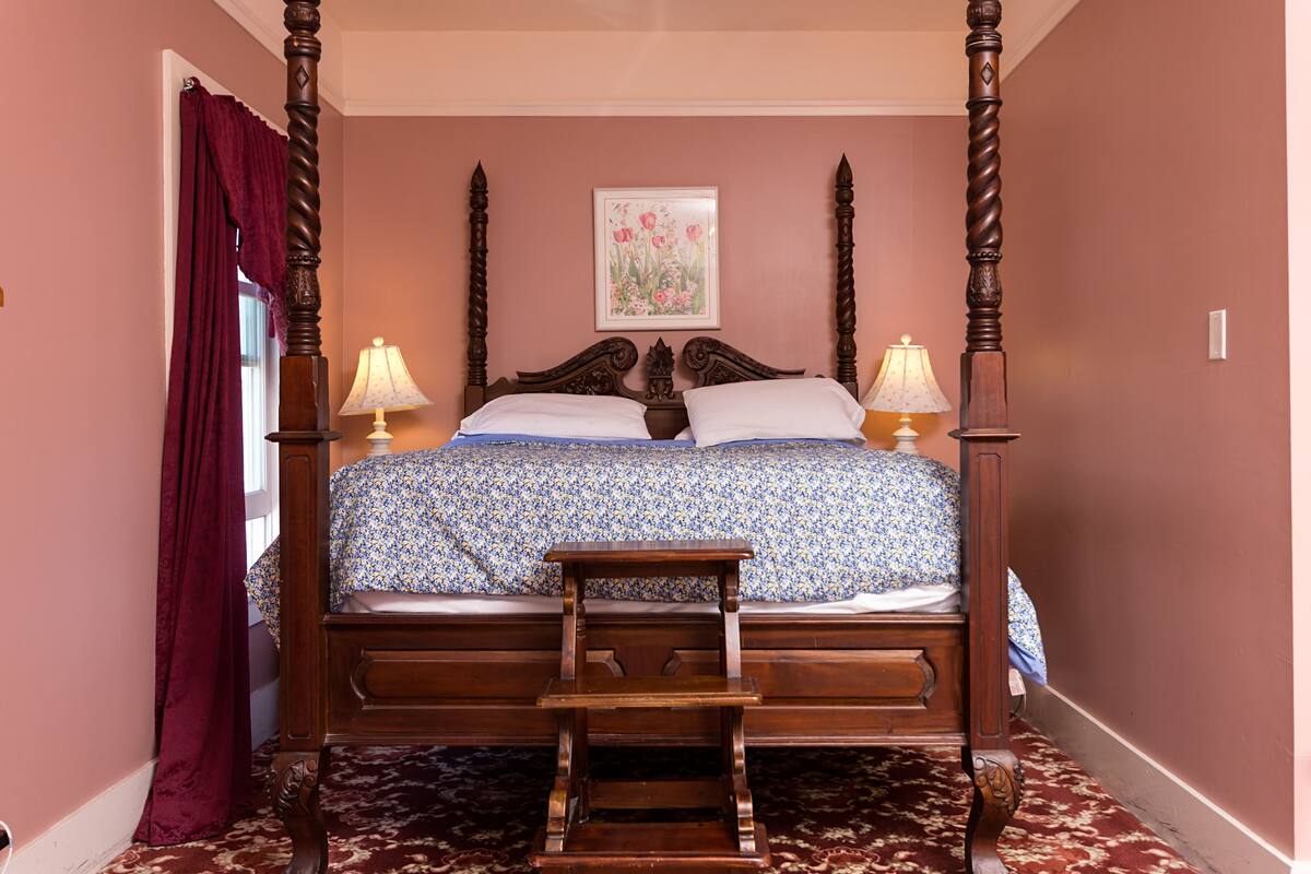 Un dormitorio con cama con dosel, dos lámparas de noche, una silla de madera y un cuadro floral en la pared.
