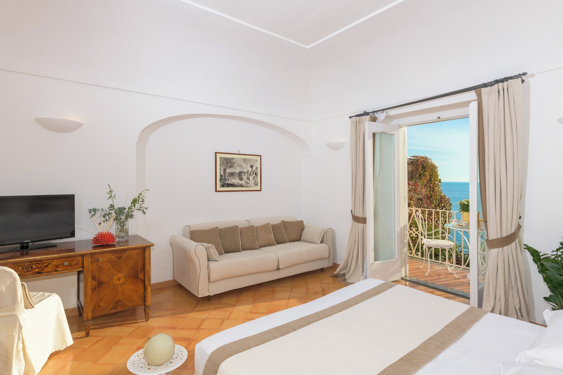 A bedroom with a bed, a TV, a sofa, a desk, and a balcony with a view of the sea.