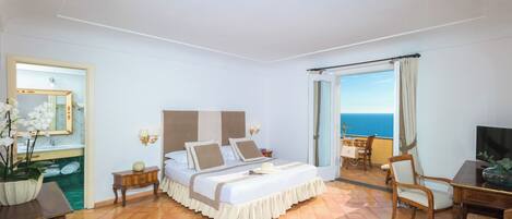 A hotel room with a bed, a desk, a chair, a TV, a bathroom with a mirror, and a view of the sea.
