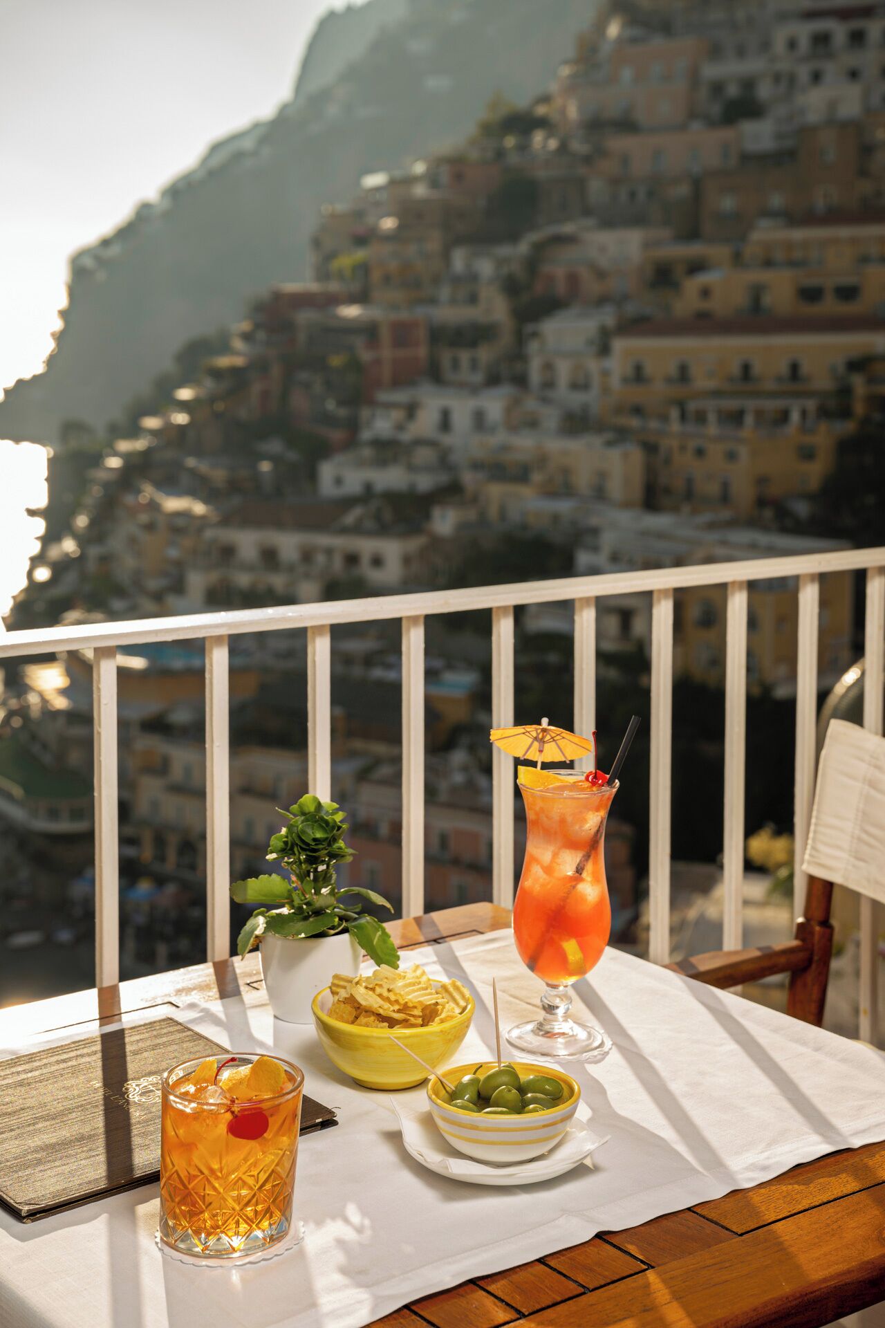 A balcony with a table set for drinks, a potted plant, and a view of a coastal town.