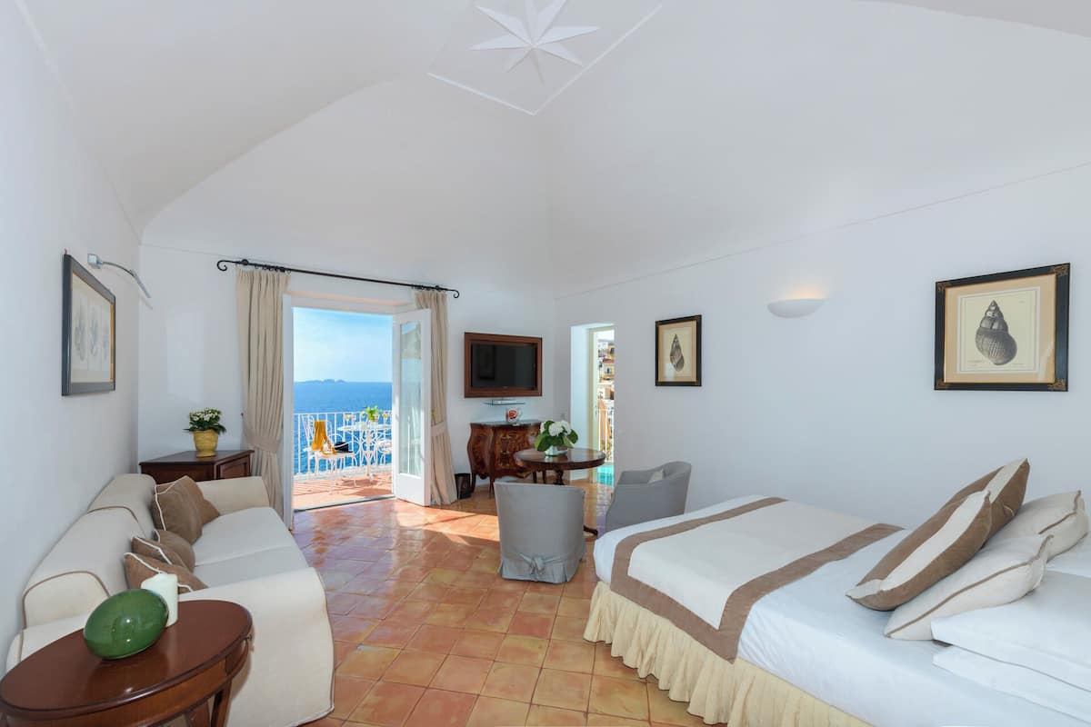 A hotel room with a bed, a sofa, a TV, a balcony with a view of the sea, and a dining area.
