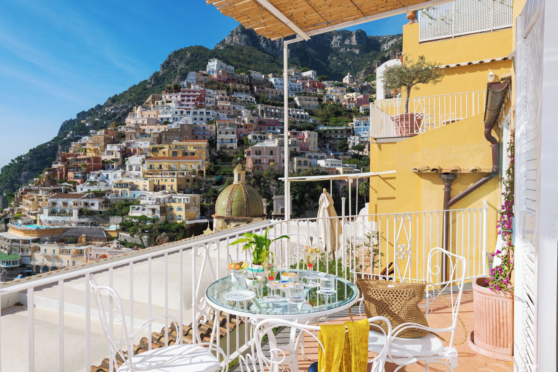 Un balcón con una mesa de vidrio y sillas blancas, con vistas a un pueblo en la ladera con edificios coloridos y una montaña al fondo.