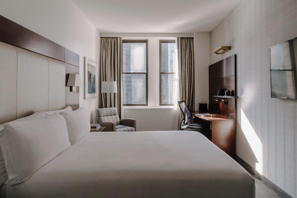 Ein Hotelzimmer mit einem großen Bett, einem Schreibtisch, einem Sessel, einem Fernseher und einem Fenster mit Vorhängen.