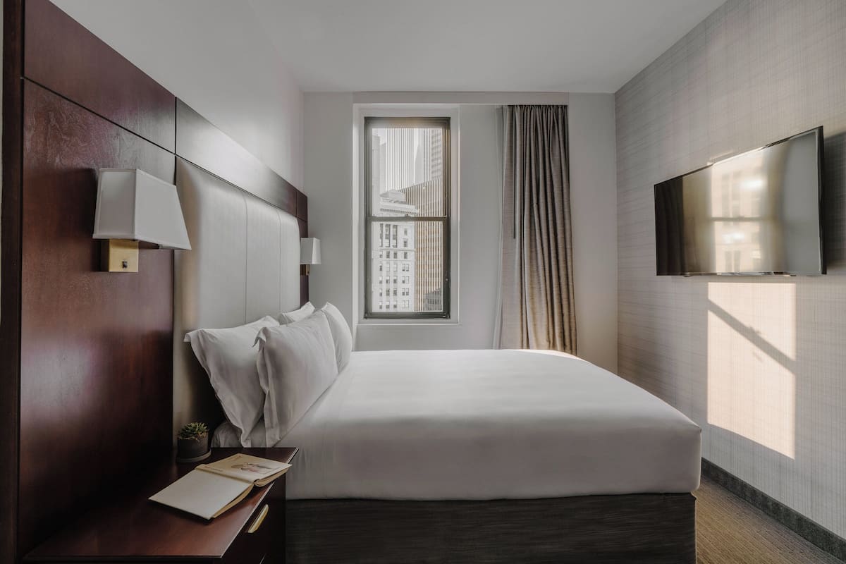 Ein modernes Hotelzimmer mit einem großen Bett, einem flachbildfernseher an der Wand, einem Nachttisch mit Lampe und einem fenster mit Vorhängen.