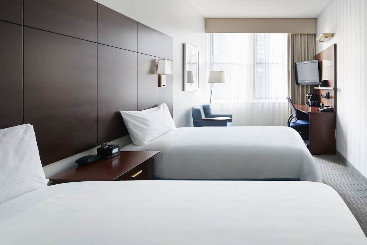 Ein Hotelzimmer mit zwei Betten, einem Schreibtisch mit Telefon, einem Sessel, einem Fernseher und einem Fenster mit Vorhängen.