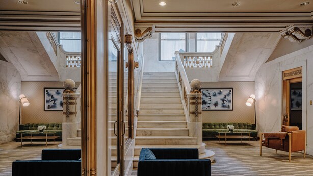 Eine prachtvolle Treppe in der Hotelhalle mit Marmorwänden, einem gemusterten Teppich und Sitzbereichen mit blau-braunen Polsterstühlen.