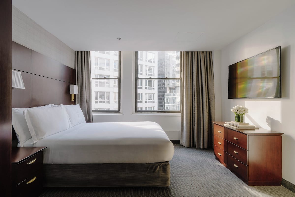 Ein Hotelzimmer mit einem großen Bett, einem Schminktisch, einem Flachbildfernseher und einem Fenster mit Vorhängen.
