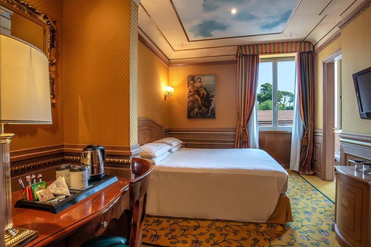 Ein Hotelzimmer mit Bett, Schreibtisch mit Kaffeemaschine, einem Bild an der Wand und einem Fenster mit Vorhängen.