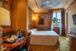 A hotel room with a bed, a desk with a coffee maker, a painting on the wall, and a window with curtains.