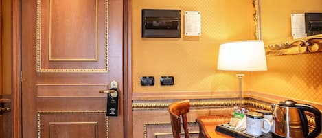 Quarto de hotel com porta de madeira, uma mesa com abajur, uma cadeira e uma cafeteira.