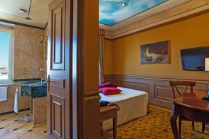 Quarto de hotel com banheiro de mármore, cama com colcha branca e almofadas vermelhas, um quadro na parede e uma televisão.
