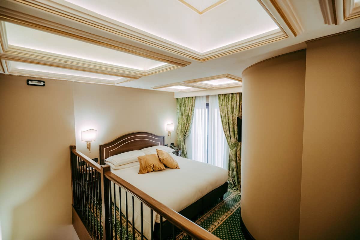 Ein Hotelzimmer mit einem großen Bett, einem Holz-Kopfteil, einem Nachttisch, einer Lampe und Blick ins Freie durch Vorhänge.
