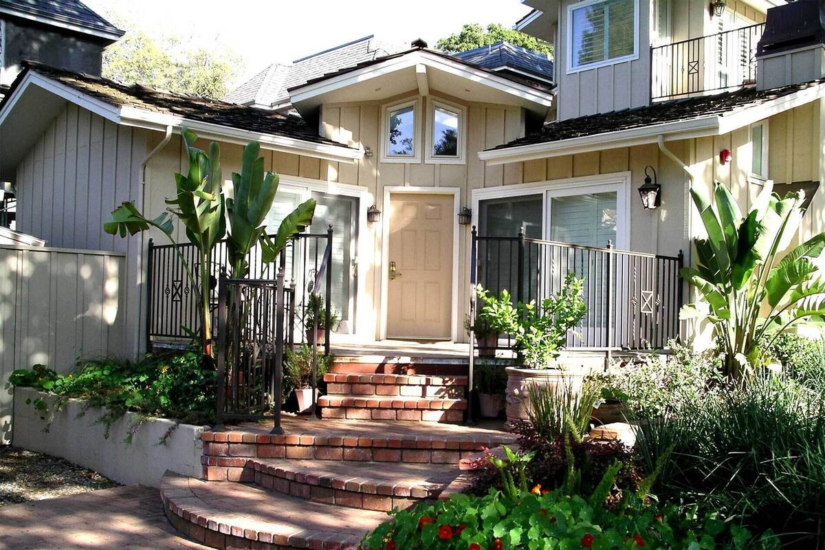 Une maison à deux étages avec un porche à l’avant, une entrée en briques et un petit jardin agrémenté de diverses plantes et fleurs.