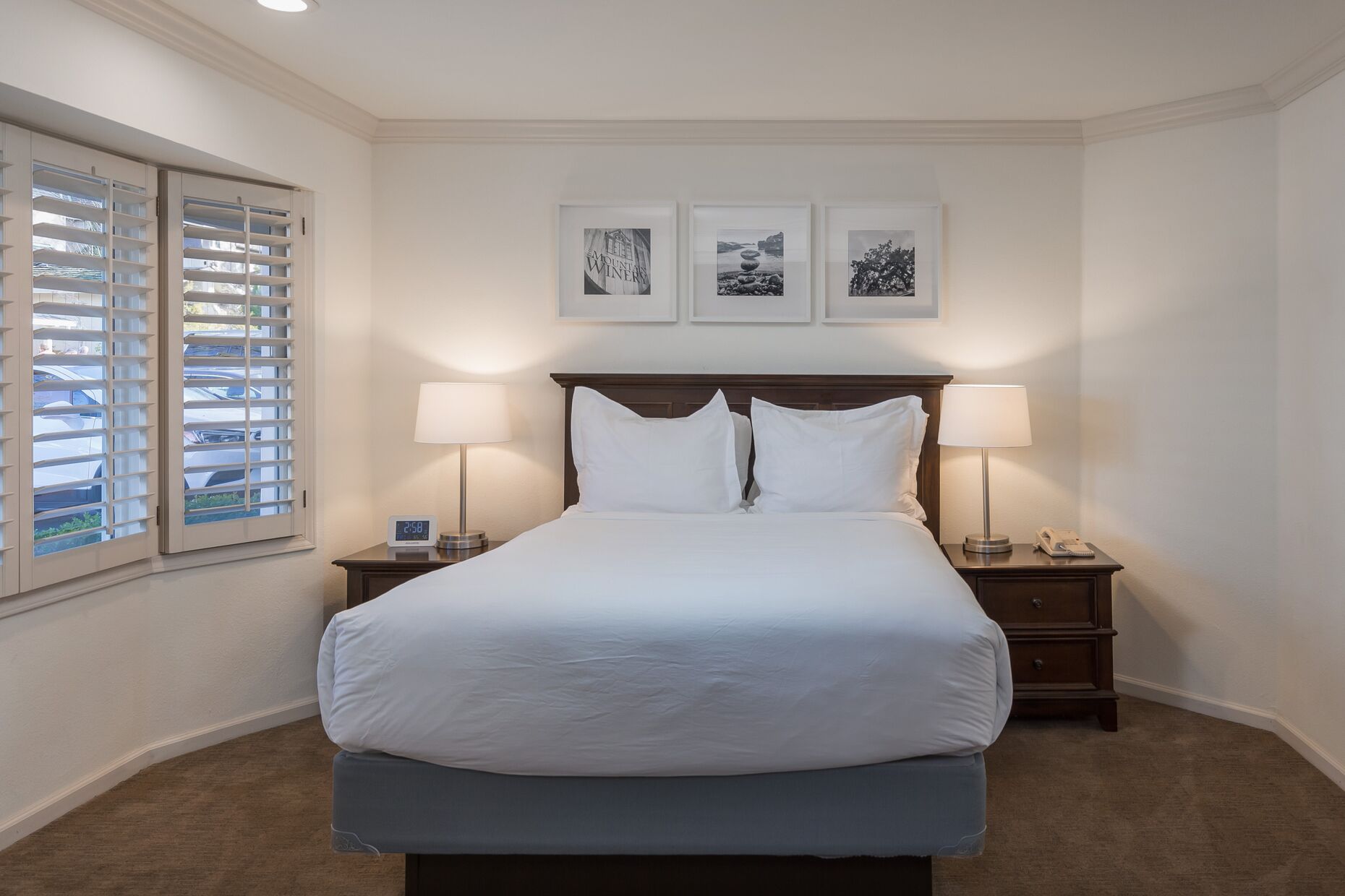 A neatly made bed in a hotel room with a wooden headboard, two bedside lamps, a clock, and three framed pictures on the wall.